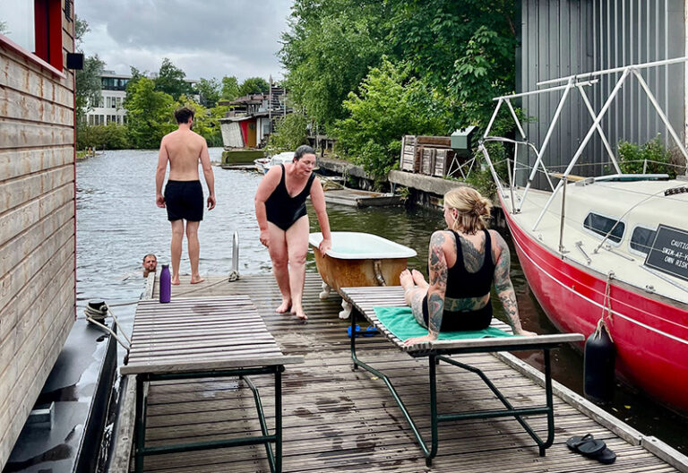 Vier Menschen verbringen Zeit an einem hölzernen Steg an einem Kanal. Ein Mann befindet sich im Wasser, während eine Frau im Badeanzug gerade aus dem Wasser steigt und sich an einer alten, freistehenden Badewanne abstützt. Eine andere tätowierte Frau sitzt auf einer Liege und schaut ihr zu. Im Hintergrund sind Hausboote und grüne Bäume zu sehen. Die Szene wirkt entspannt, urban und gemeinschaftlich, bei bewölktem Himmel.