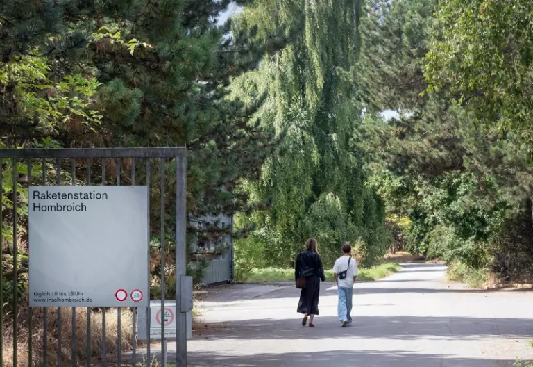 Das Bild zeigt den Eingang der Raketenstation Hombroich bei Neuss. Zwei Frauen gehen durch das Eingangstor.